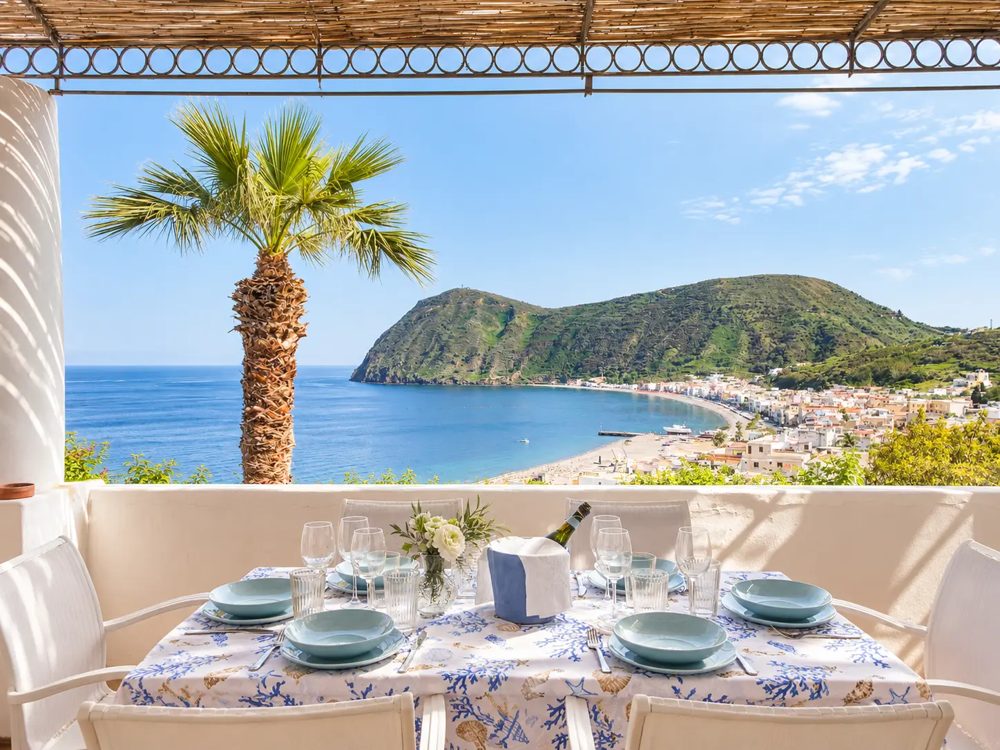 La tavola in terrazza sotto l’incannucciato, con la palma e il mare